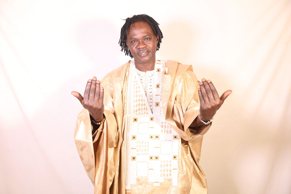 Baaba Maal wearing a gold robe stands against a cream-coloured background, as he beckons the viewer in with his hands.